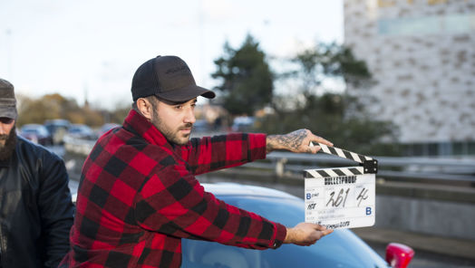A man wearing a red checked shirt and baseball cap holds a clapper board in front of a sports car