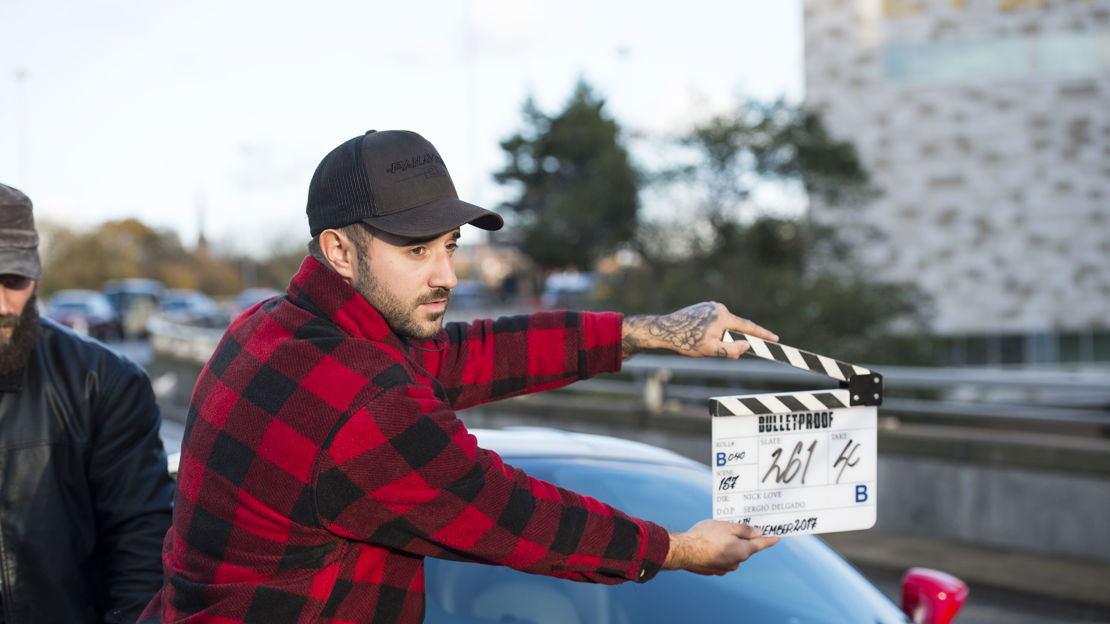 A man wearing a red checked shirt and baseball cap holds a clapper board in front of a sports car