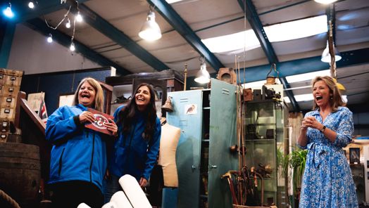 Three women stand laughing in a room filled with antiques on the set of Bargain Hunt
