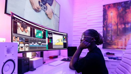 A person sits at a desk in front of a large monitor. On the screen are images in a video editing software. To the left of the screen is a speaker.