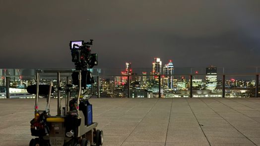 A camera stands on a moving platform on a roof overlooking a nighttime cityscape