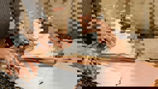 Two people sit at a desk with laptops and notebooks between them