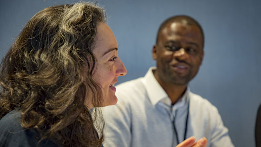 Woman in the foreground speaking and motioning with hands and a man in the background watching