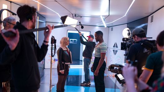 On the set of Doctor Who, Ncuti Gatwa as the 15th Doctor stands facing Millie Gibson as Ruby Sunday surrounded by production crew