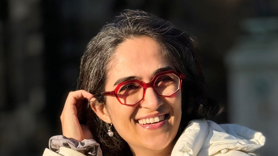 Elahe Esmaili stands smiling in a white coat with her arm raised to push back hair