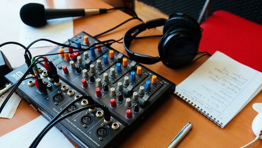A picture of an audio mixer on a desk. Also on the desk is a notebook and pen, a microphone and a pair of headphones.