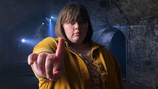 Flo Taylor stands facing the camera with arm held out and her index finger raised