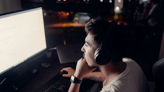 A young man wearing headphones sits in front of a laptop in a dark room in front of a window at night