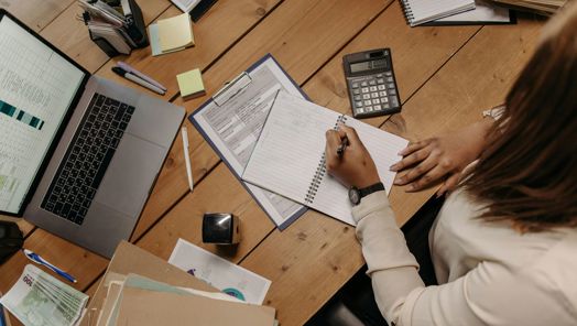 Woman sitting at a desk with an open laptop, folders and a calculator, writing into a notebook
