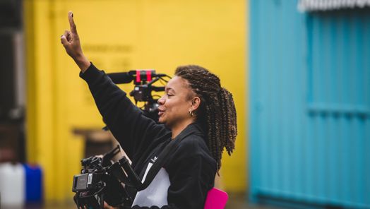 A woman stands on location holding a camera with her arm in the air