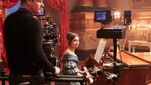An actress sits at a piano on the set of a period drama. In the foreground, a camera operator stands monitoring camera equipment.